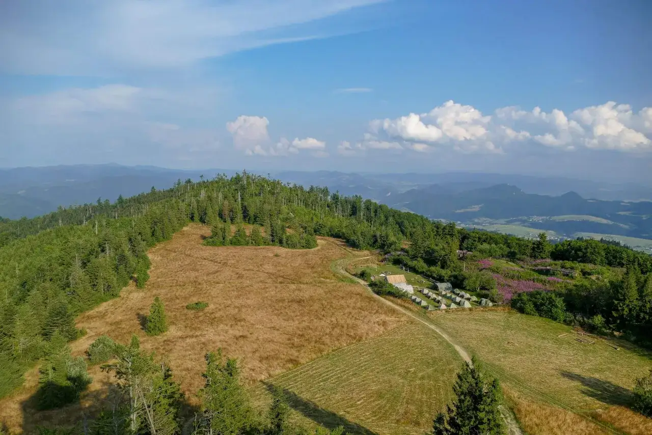Widok z lotu ptaka na g&oacute;rski krajobraz. Widać zalesione wzg&oacute;rza, pola i rozległą panoramę. Na jednym ze zboczy znajduje się ob&oacute;z z namiotami, a obok niego kręta lubań trasa.