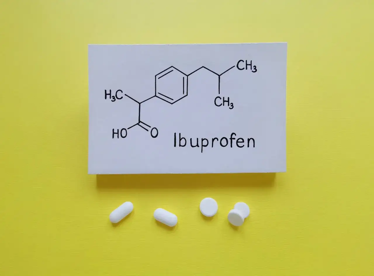 Wzór chemiczny ibuprofenu, substancji czynnej leków przeciwbólowych i przeciwzapalnych, obok białych tabletek.
