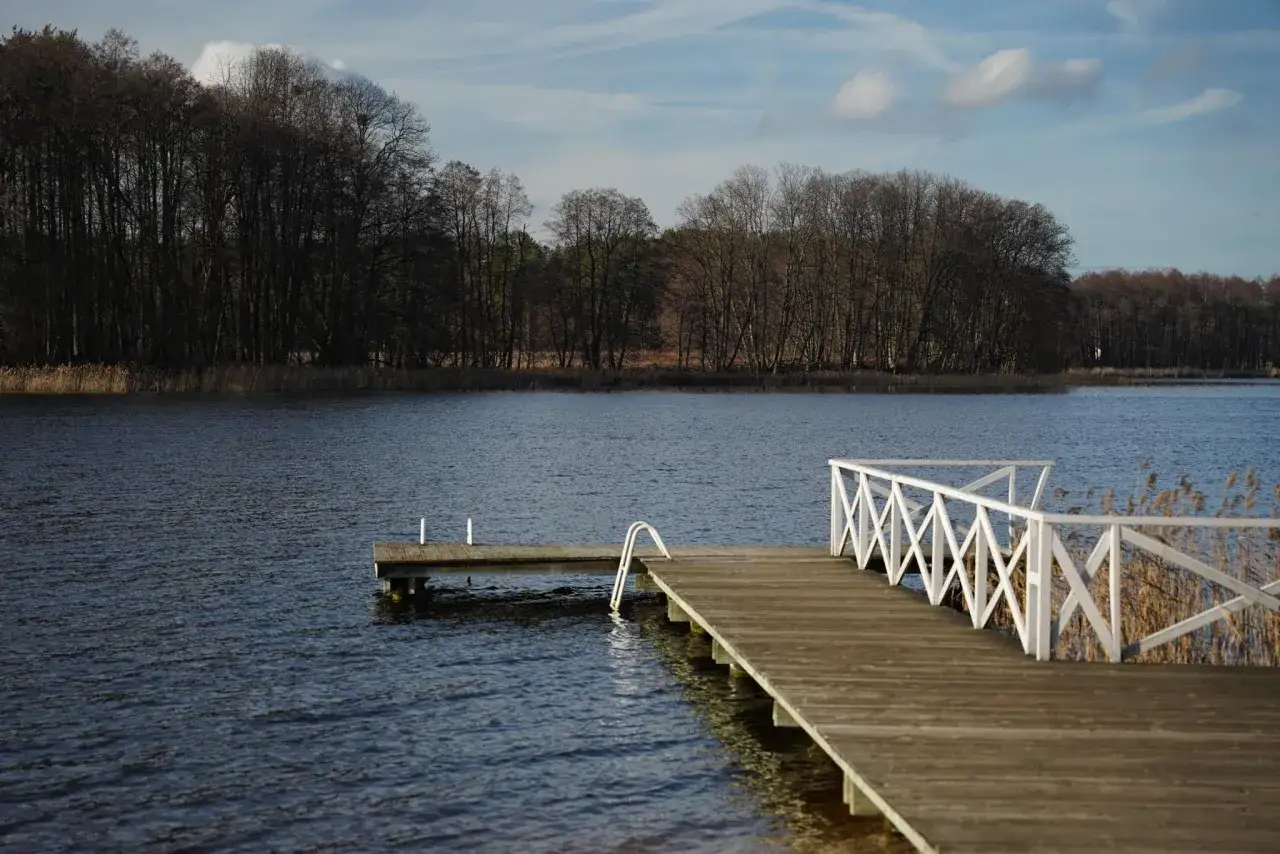 Drewniane molo z białą balustradą nad spokojnym jeziorem wejsunek. W oddali las.