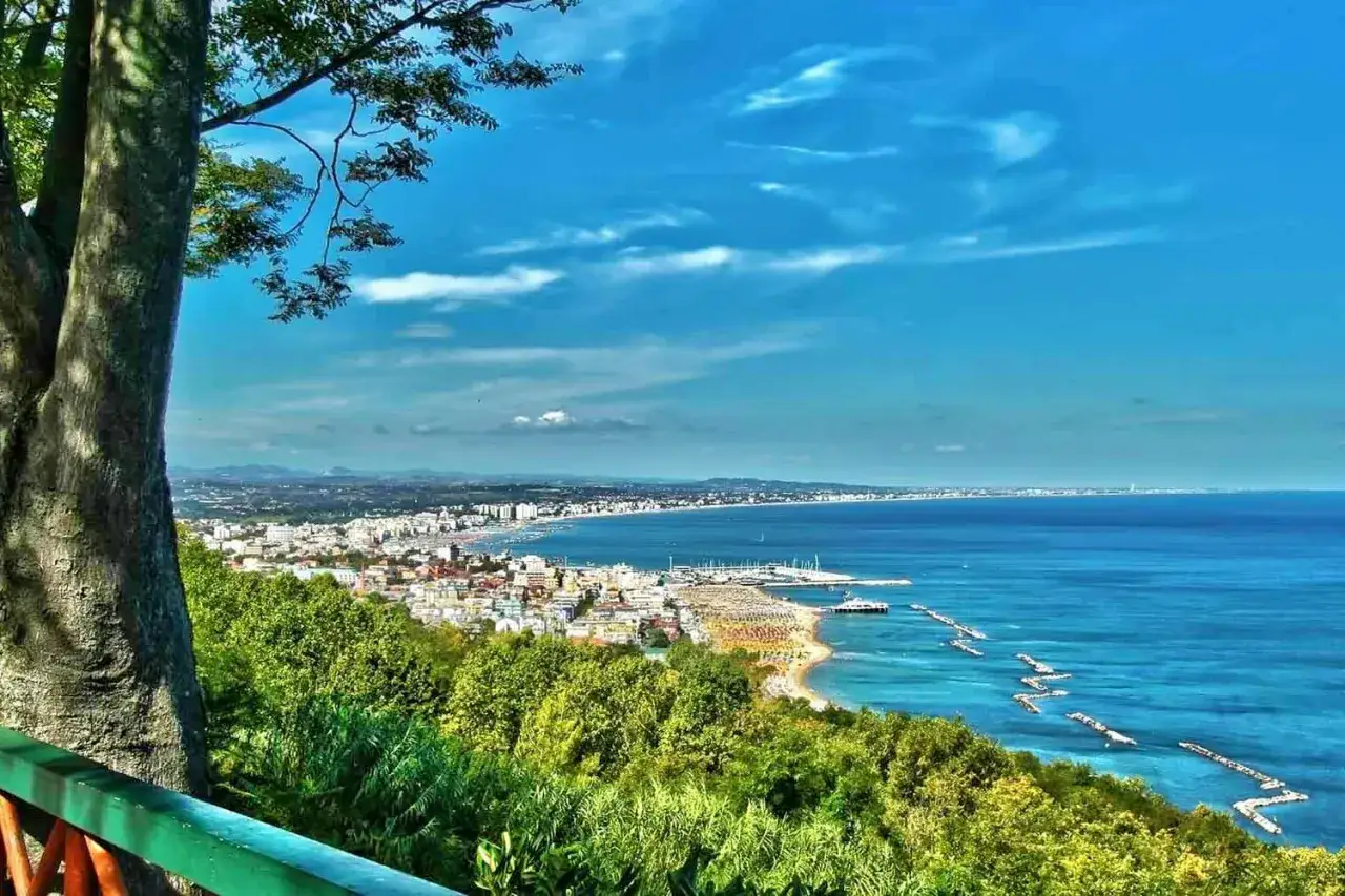 Vista panoramica della costa di Rimini con mare azzurro, spiaggia affollata e verdeggianti colline.