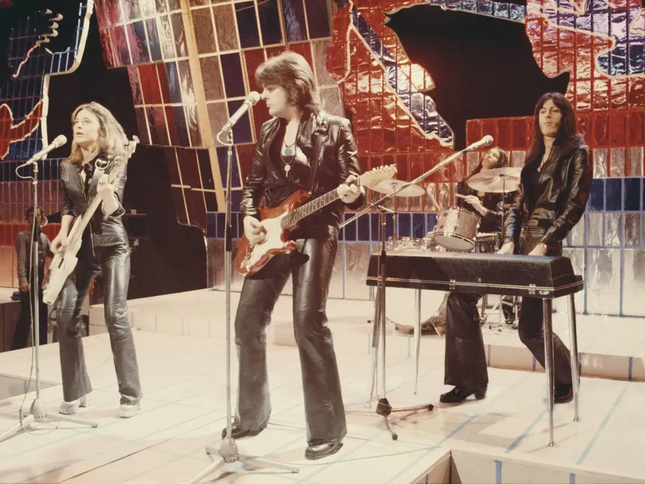Zespół grający na scenie, w tym Suzi Quatro, wykonuje swoje piosenki w skórzanych strojach.