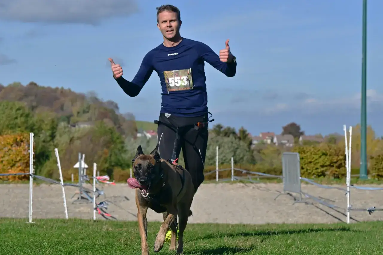 Mężczyzna z psem rasy malinois biegnie w zawodach psich sport&oacute;w. Oboje zadowoleni, pokazują kciuki w g&oacute;rę.