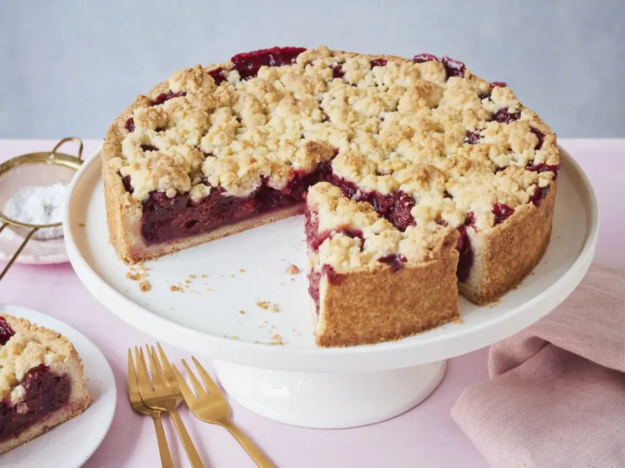 Ein saftiger Kirschkuchen mit knusprigen Streuseln auf einem weißen Tortenständer. Ein Stück ist bereits herausgeschnitten.