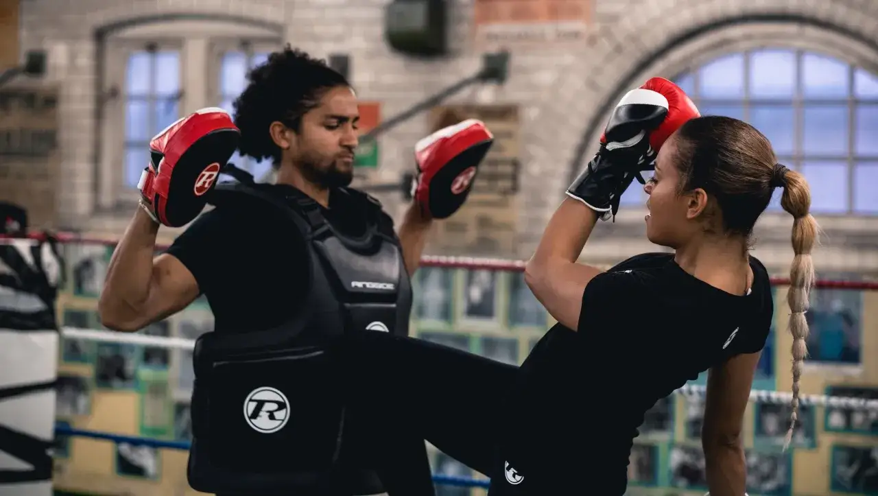 A man in a protective vest holds pads for sparing boxing as a woman in gloves delivers a powerful kick.