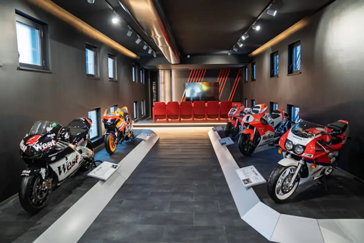 W nowoczesnym muzeum motocykli prezentowane są wyścigowe maszyny. Wnętrze z czerwonymi siedzeniami i ekranem tworzy atmosferę toru wyścigowego.