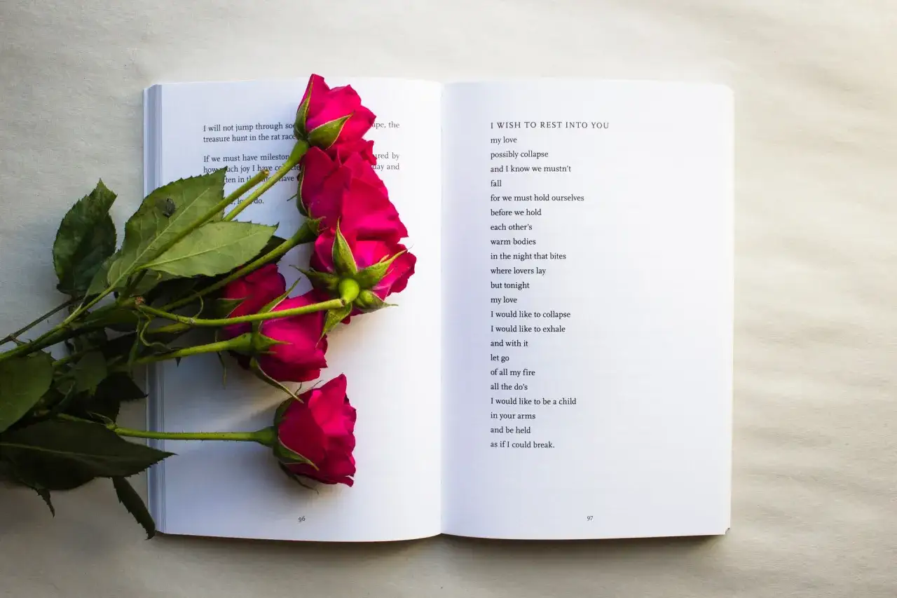 Krótkie wiersze o miłości, ozdobione bukietem róż. Na otwartej książce widać poezję, a obok niej leżą ciemnoróżowe róże.