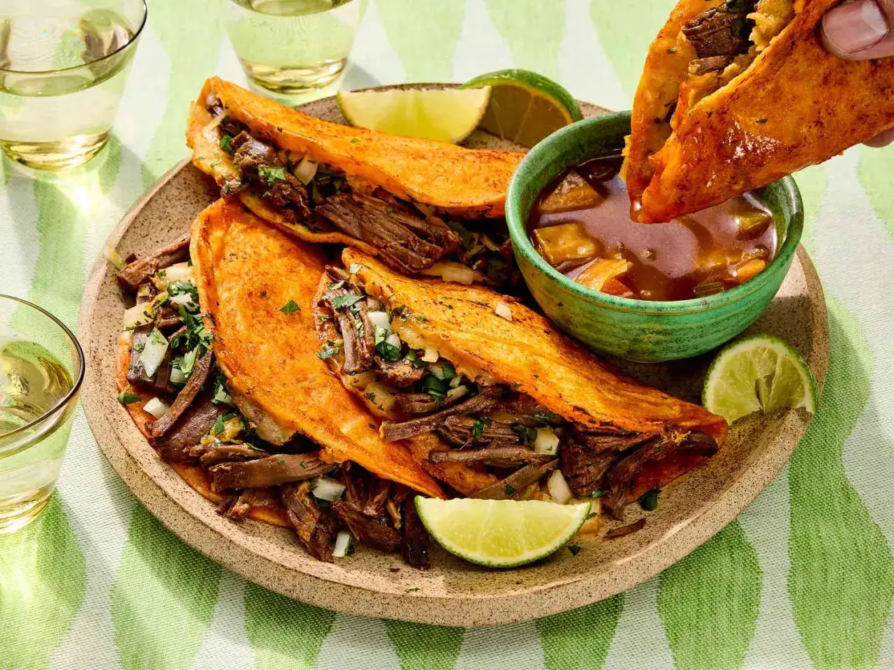 Saftige birria tacos mit Rindfleisch, Zwiebeln und Koriander, zum Dippen in Consomé. Dazu Limettenspalten und Gläser mit hellem Getränk.