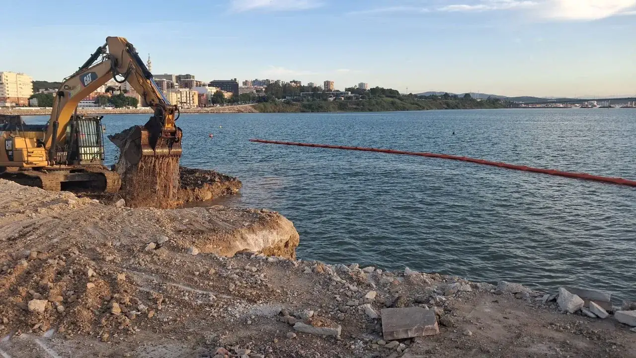 Excavadora trabajando en la costa, moviendo tierra cerca de una escollera hormigonada. En el agua, una barrera flotante naranja.