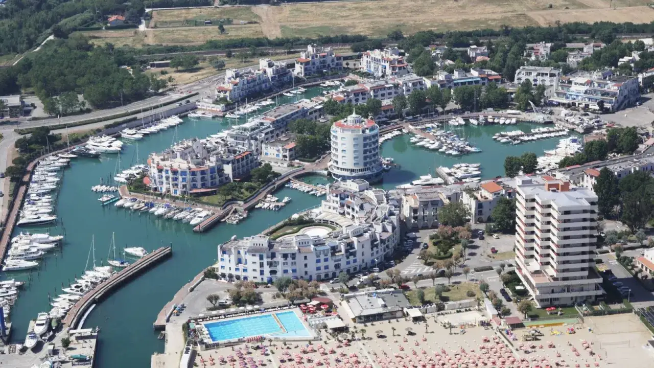 Vista aerea del porto turistico di Cattolica, con yacht ormeggiati e edifici bianchi moderni. La spiaggia e la piscina completano il quadro.