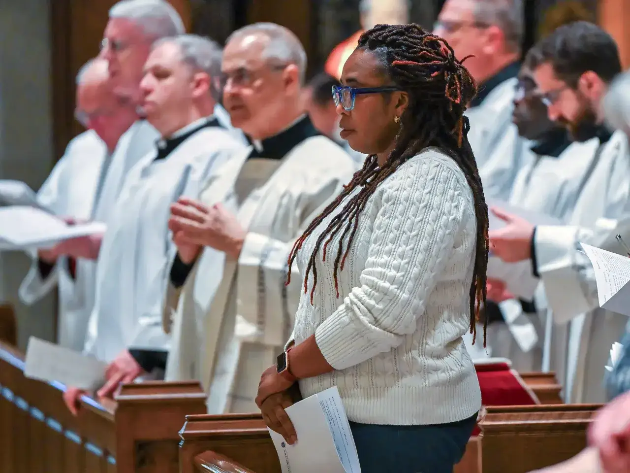 A woman with dreadlocks and glasses stands in a church, holding a program. Behind her, men in white robes participate in a religious service, perhaps an opus dai.
