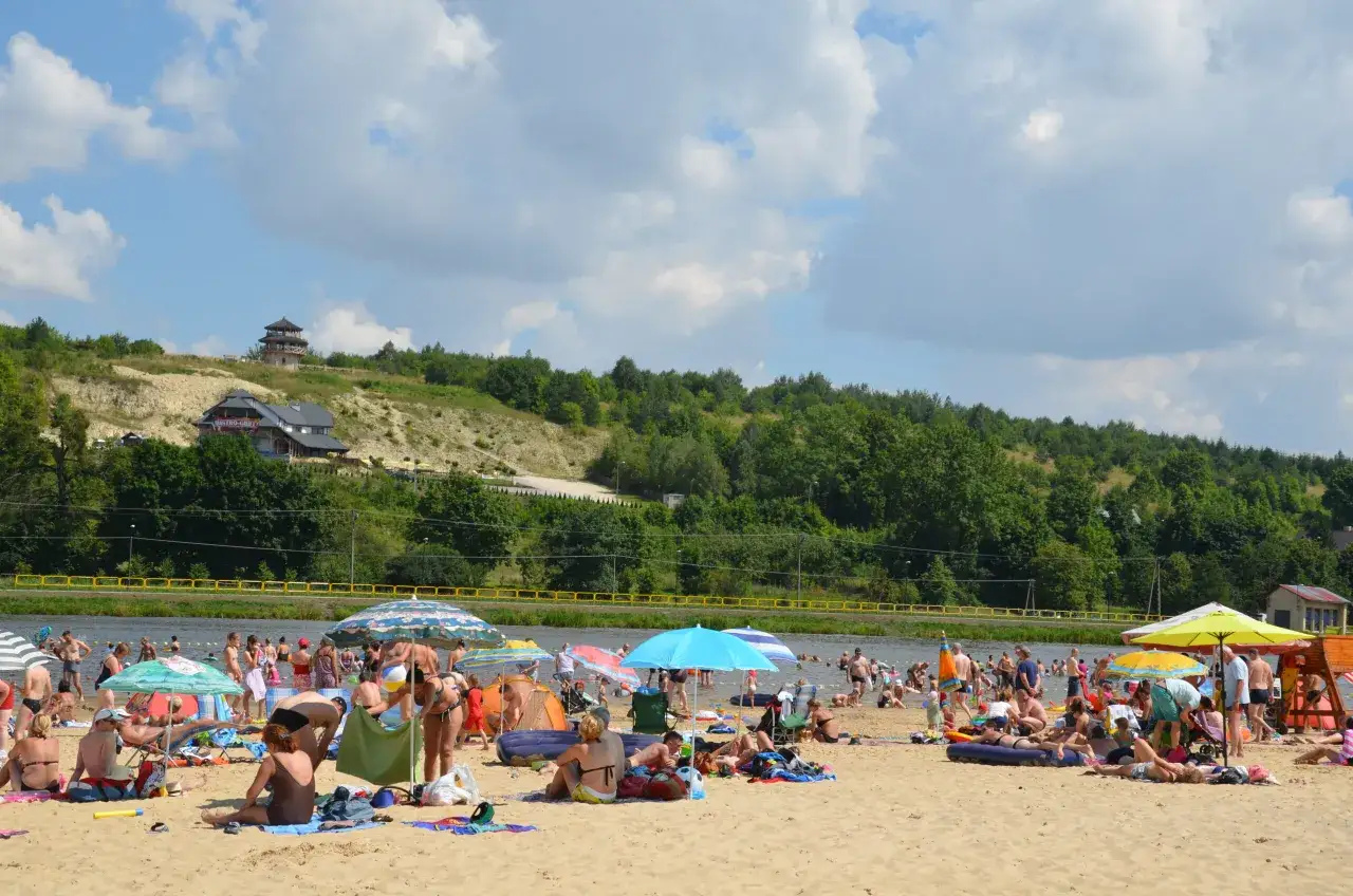 Słoneczny dzień na **Krasnobrodzie zalew**. Ludzie odpoczywają na plaży, kąpią się w wodzie, pod kolorowymi parasolami.