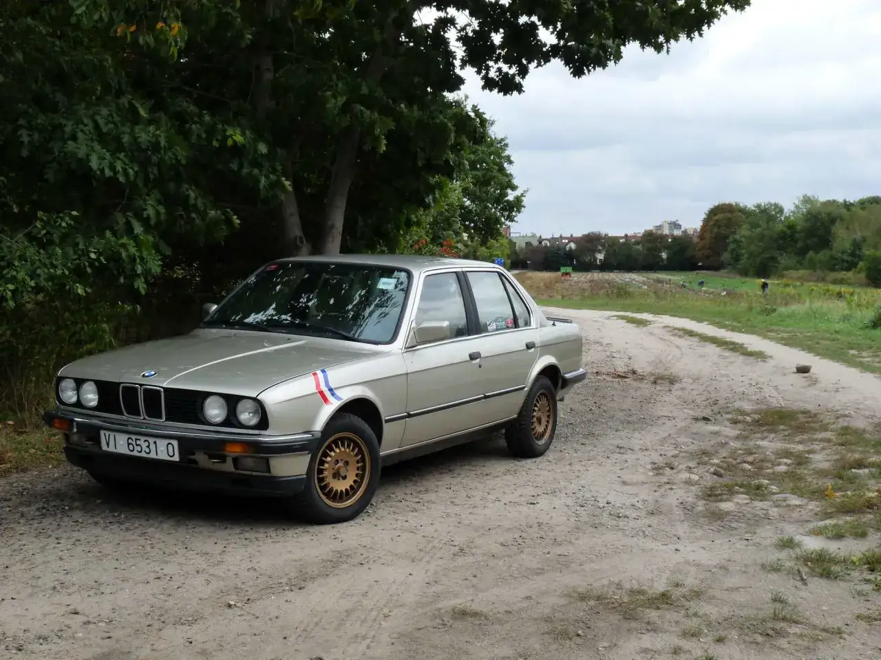 Srebrne BMW E30 coupe z francuskimi barwami na błotniku stoi na piaszczystej drodze wśród zieleni.