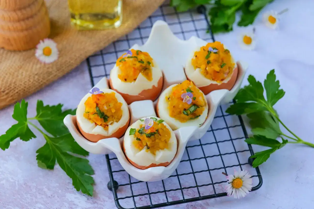 Cinq &oelig;ufs mimosa garnis d'une pur&eacute;e orang&eacute;e et d'herbes, d&eacute;cor&eacute;s de petites fleurs violettes, pr&eacute;sent&eacute;s dans un plat blanc en forme d'&oelig;uf.