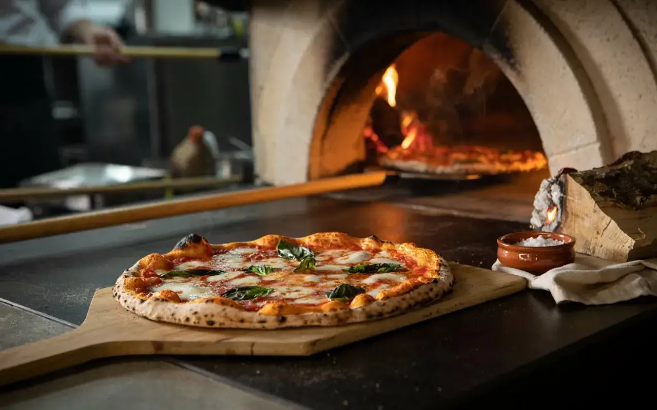 Margherita pizza na desce, pieczona w piecu opalanym drewnem. Tak właśnie powstaje pizza, której smak przenosi nas do Neapolu.