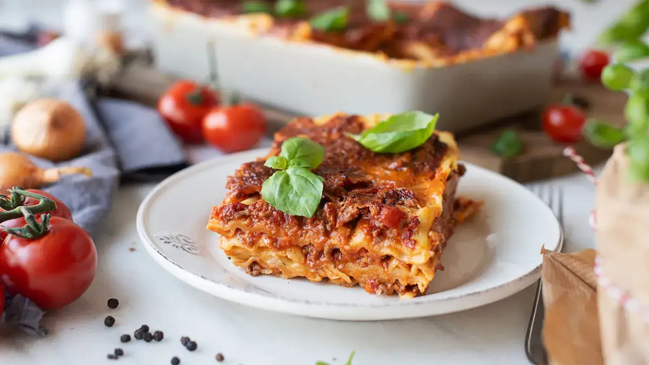 Ein St&uuml;ck Lasagne al forno mit frischem Basilikum, daneben Tomaten und Zwiebeln.