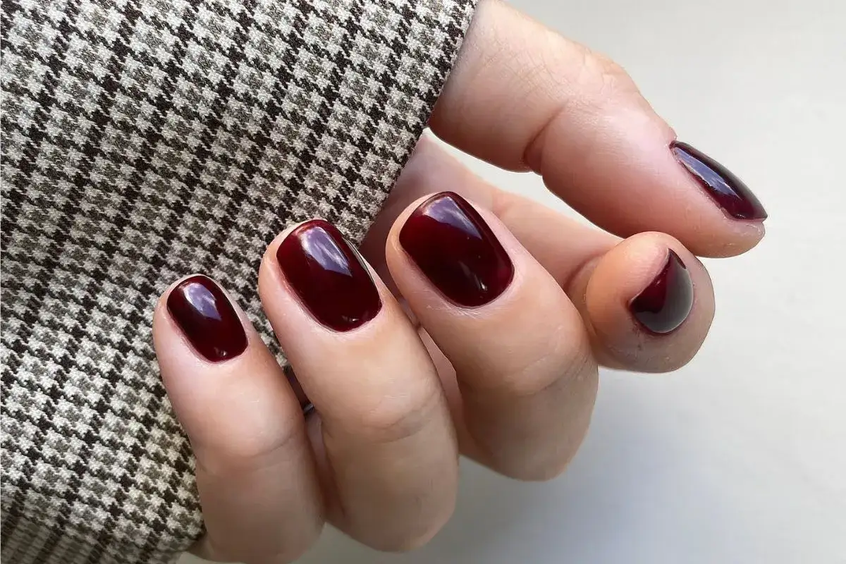 Dłoń w eleganckim żakiecie z pepitką, prezentująca modne kolory paznokci w odcieniu głębokiej wiśni.