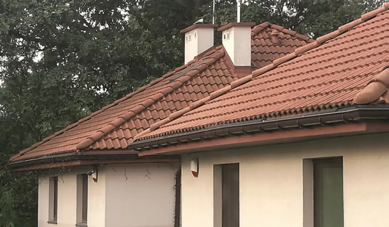 Montaż rury spustowej na dachu z dachówki ceramicznej. W tle drzewa.