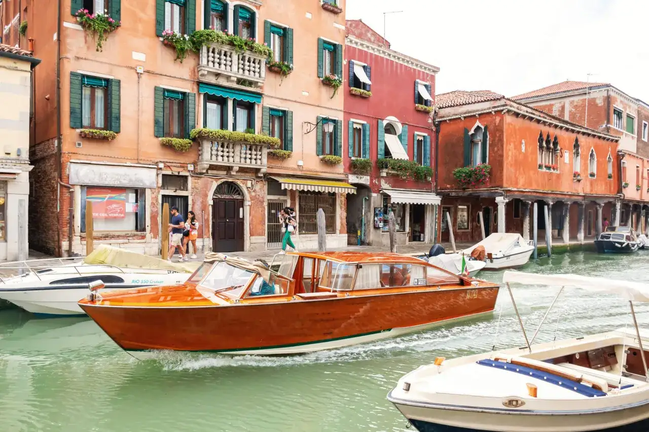En vacker träbåt glider fram på en kanal i Venedig, omgiven av färgglada hus och andra båtar.