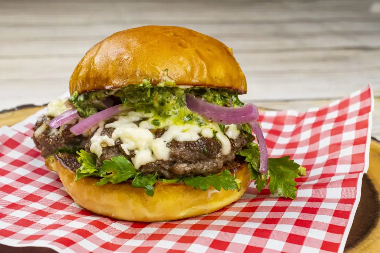 Un délicieux burger agneau avec fromage fondu, oignons rouges et sauce verte, servi sur un papier à carreaux rouges et blancs.