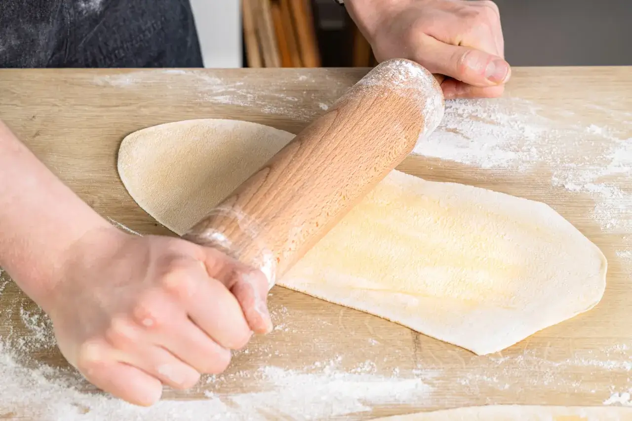 Hände rollen Teig für Maultaschen aus. Der Nudelholz verteilt das Mehl gleichmäßig auf dem Holzbrett.