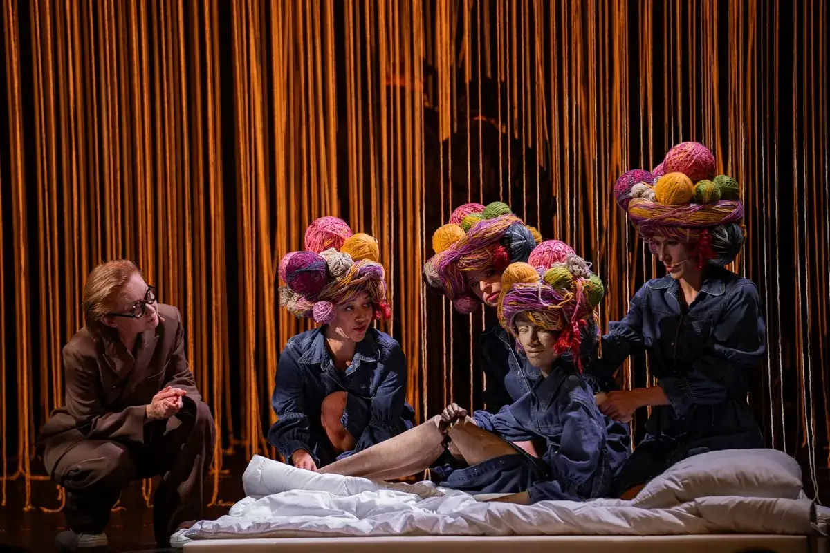 Na scenie, gdzie życie to nie teatr interpretacja, cztery postacie w barwnych perukach z włóczki i denimowych strojach. Jedna siedzi, dwie klęczą, a jedna stoi, otaczając leżącą postać.