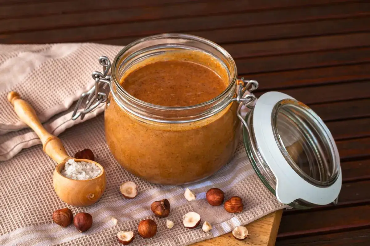 Un pot de pralin&eacute; maison onctueux, entour&eacute; de noisettes enti&egrave;res et bris&eacute;es, avec une cuill&egrave;re en bois remplie de gros sel.