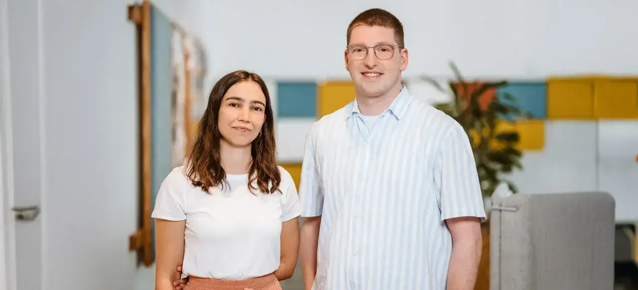 Ein junges Team, bestehend aus einer Werkstudentin Marketing und ihrem Kollegen, posiert l&auml;chelnd im B&uuml;ro.