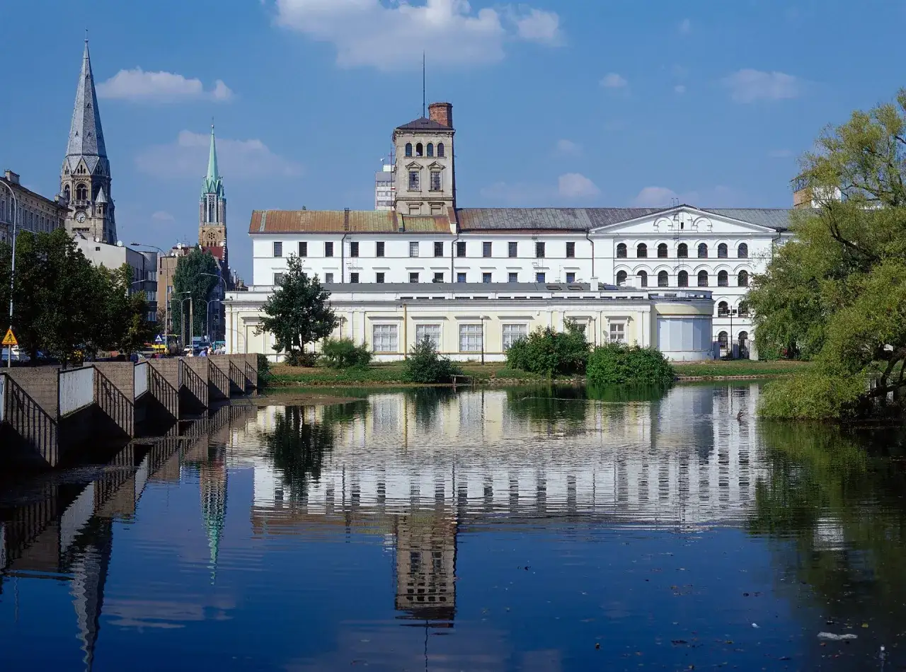 Zabytkowe budynki fabryczne w mieście w województwie łódzkim nad Wartą, odbijające się w spokojnej tafli wody.
