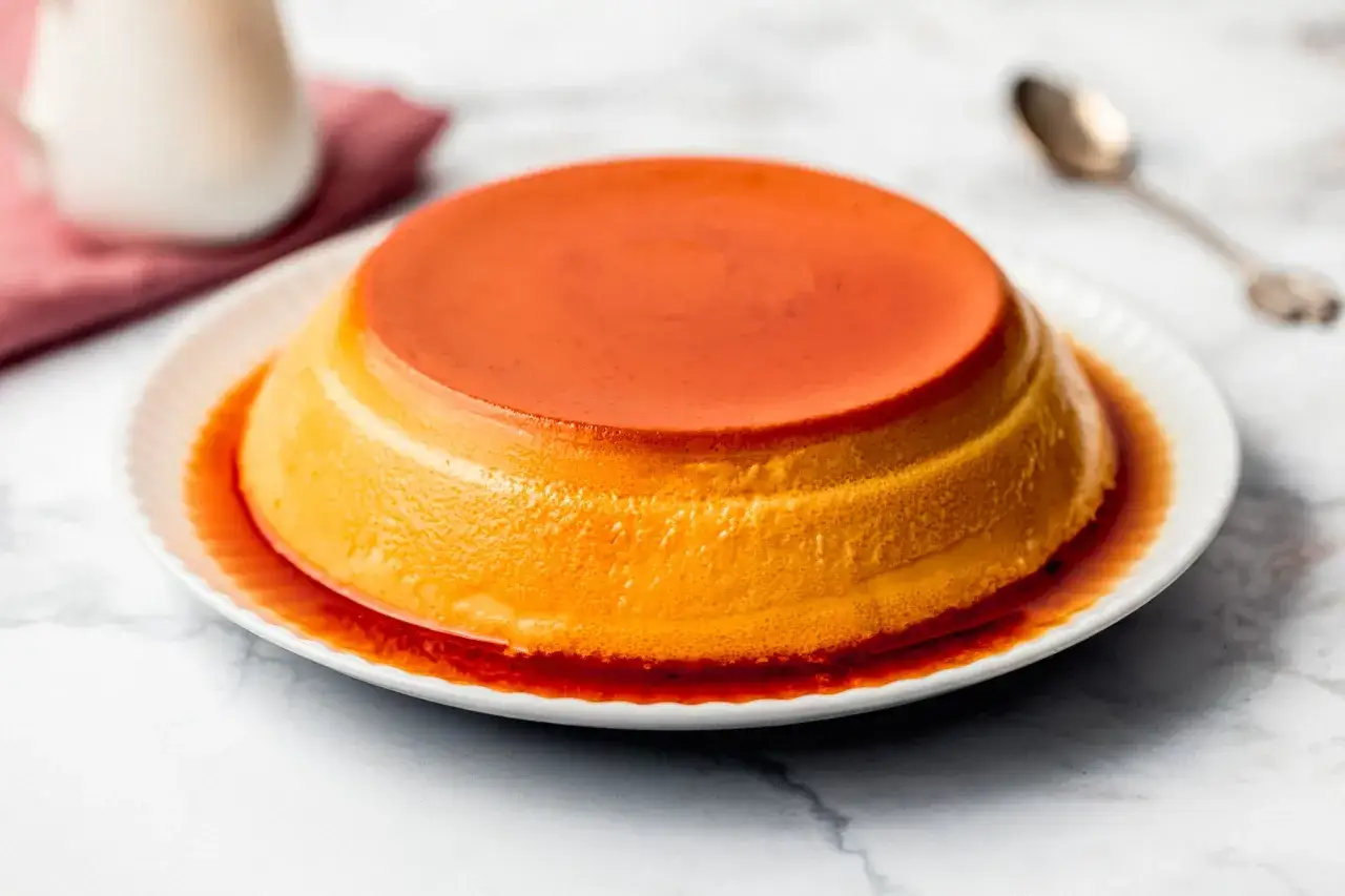 Delizioso creme caramel dorato, con caramello liquido che cola sul bordo del piatto bianco.