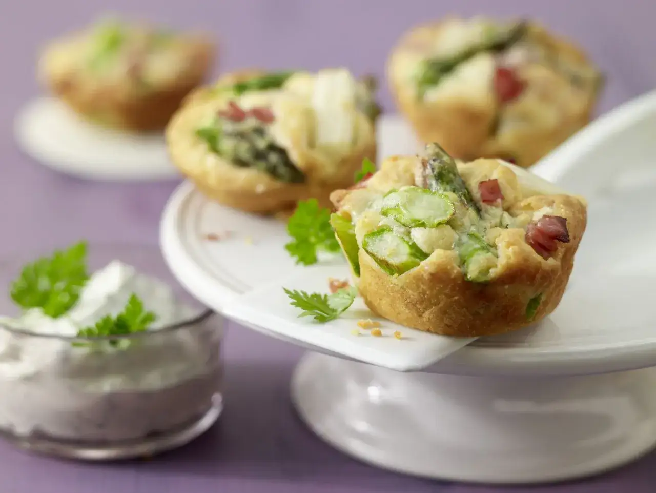Herzhafte Muffins mit Spargel und Speck, garniert mit Petersilie, neben einer Schale Dip.