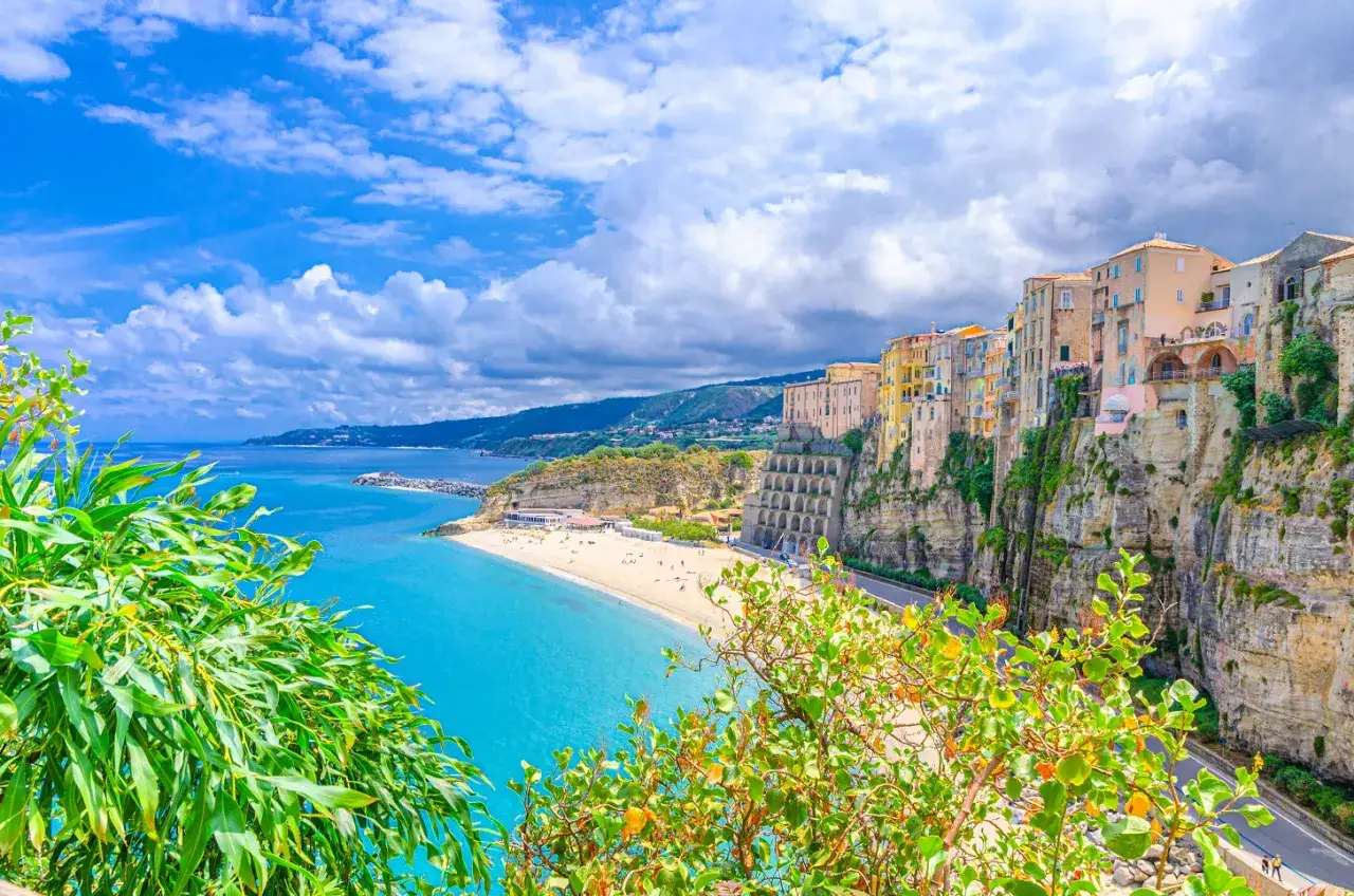 Case colorate a picco sul mare turchese a Tropea, Vibo Valentia. Spiaggia dorata e cielo azzurro con nuvole bianche.