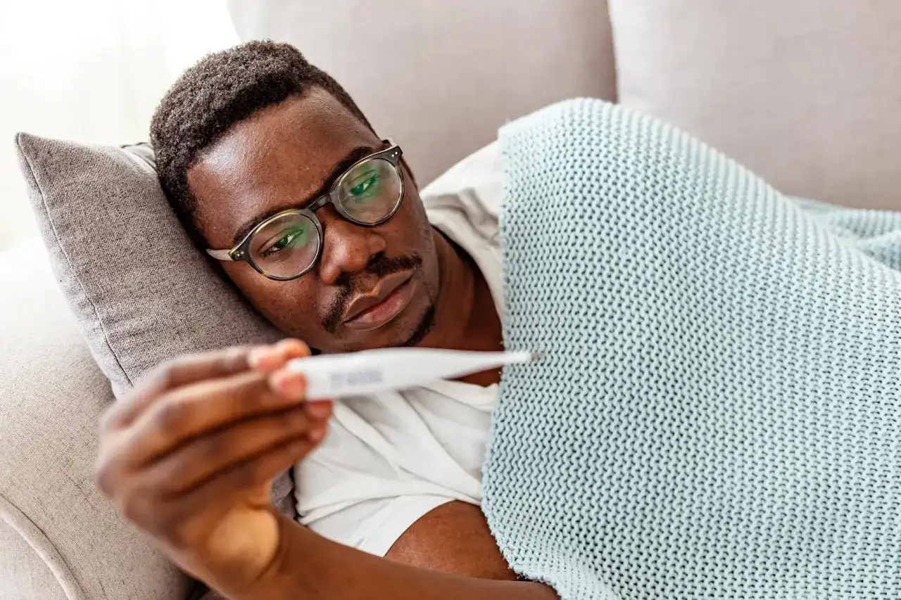 Man med glasögon ligger under en filt och mäter feber. Kan man få feber av stress?