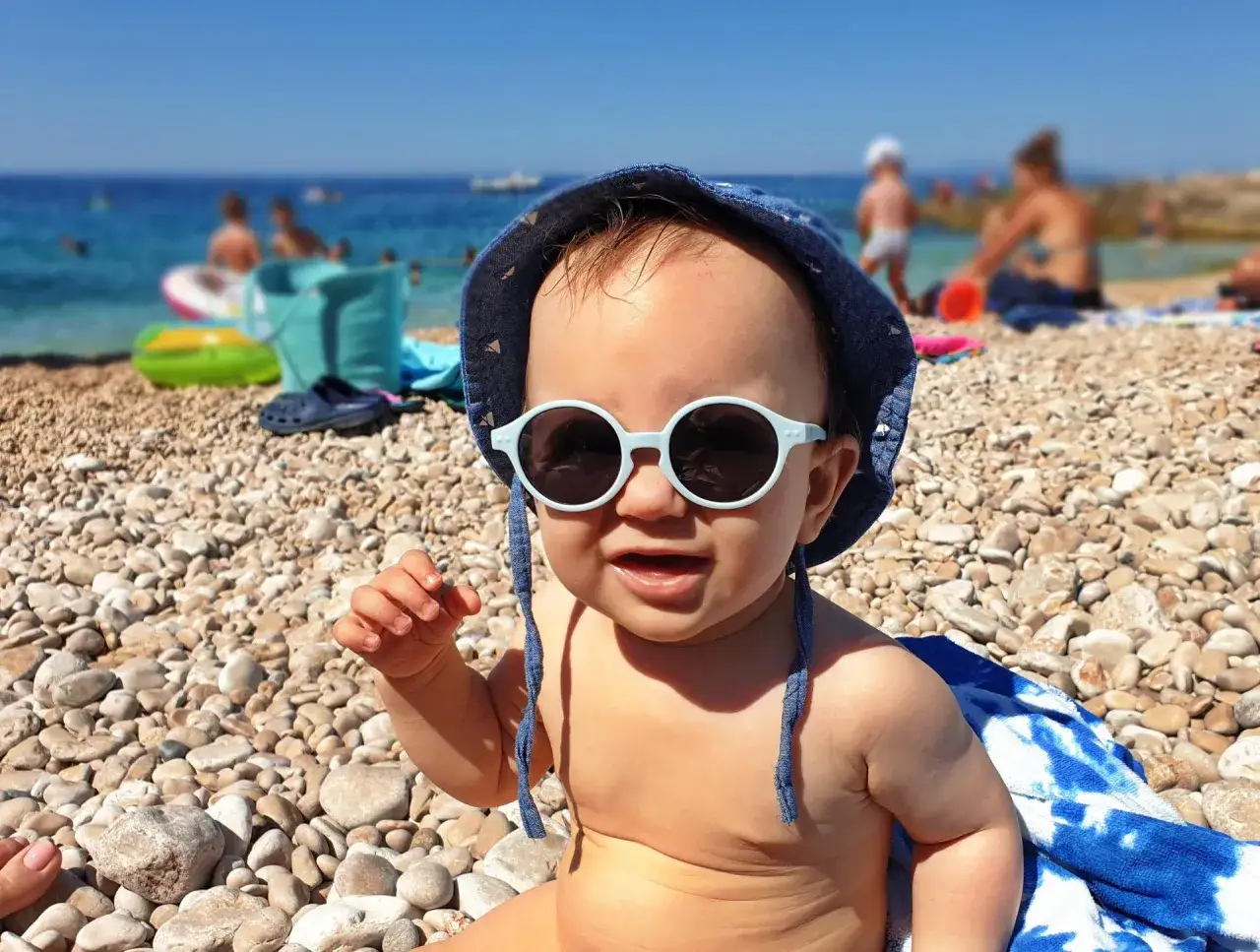 Urocze dziecko w okularach przeciwsłonecznych i kapeluszu na kamienistej plaży w Chorwacji. Idealne rzeczy, co zabrać na plażę w Chorwacji: ręcznik, zabawki do piasku i krem z filtrem.