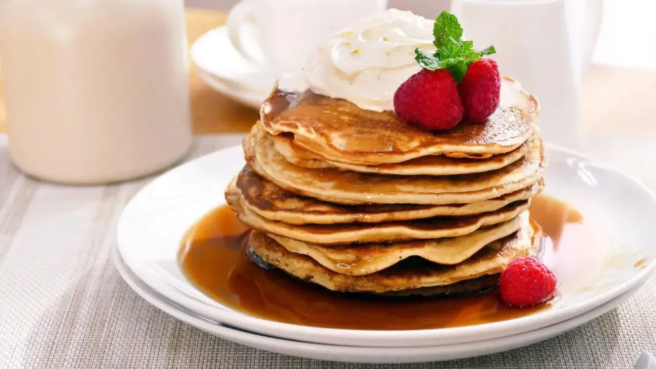 Una pila de tortitas americanas cubiertas con crema batida, frambuesas y sirope.