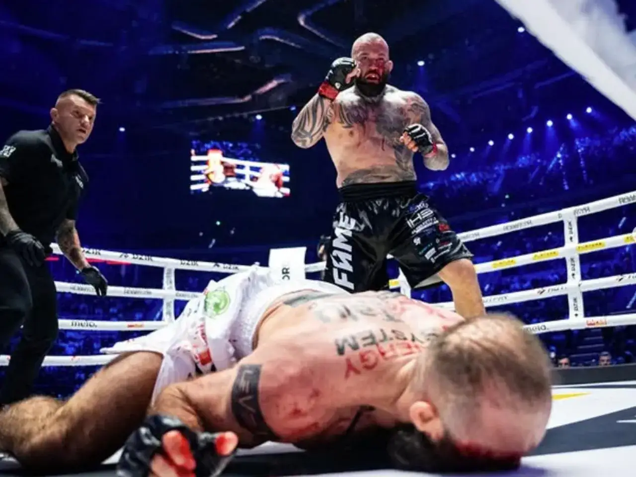 Zwycięzca gali FAME MMA triumfuje nad pokonanym rywalem. Sędzia czuwa nad przebiegiem walki.