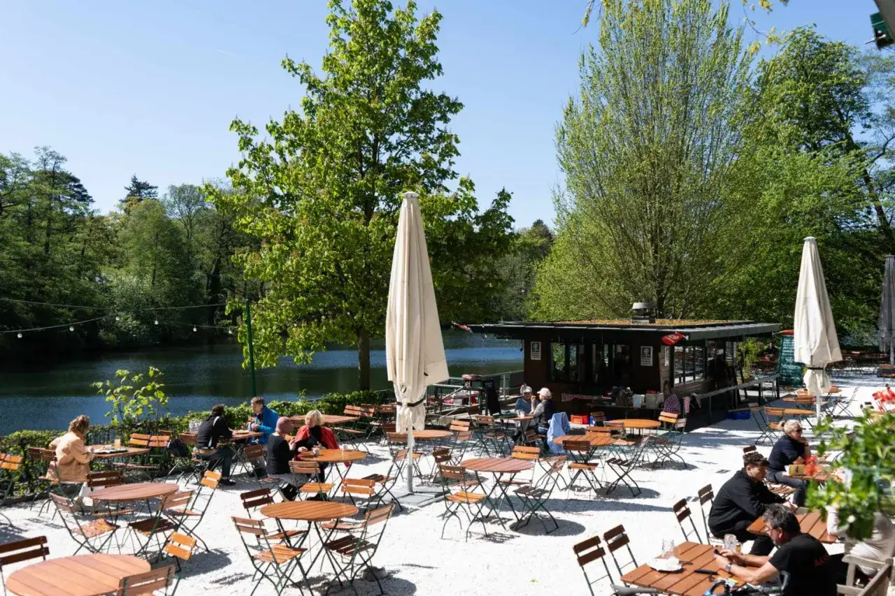 Gem&uuml;tliche Biergartenatmosph&auml;re am Schlachtensee Berlin. G&auml;ste genie&szlig;en das sonnige Wetter an Holztischen mit Blick auf den See und die gr&uuml;ne Natur.
