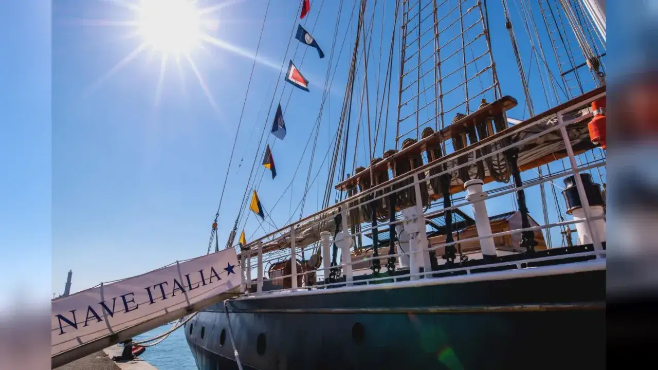 La nave Italia, con le sue vele spiegate e le bandiere colorate, si staglia contro un cielo azzurro e soleggiato.