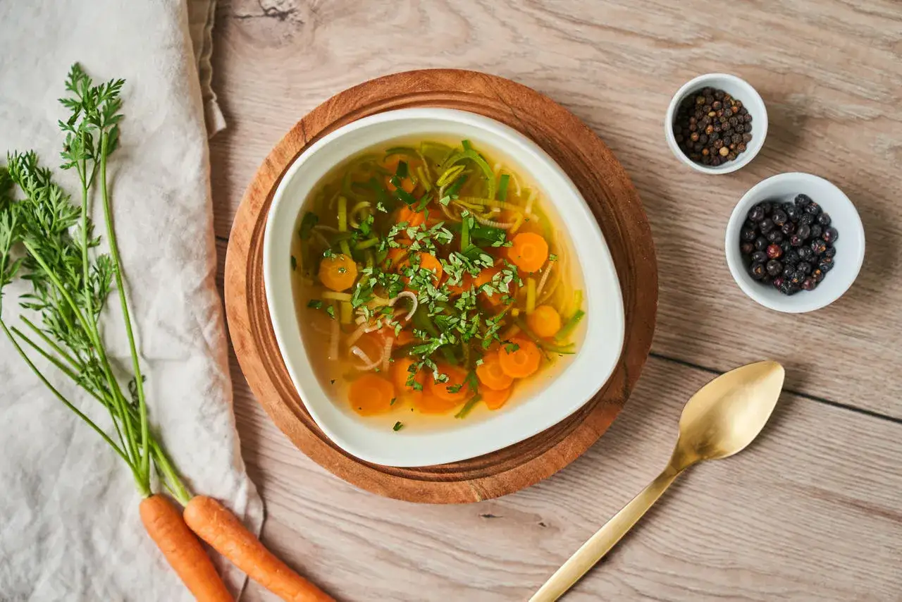 Un bol de bouillon de légume réconfortant, garni de carottes et de fines herbes. À côté, des carottes fraîches et des épices pour assaisonner.