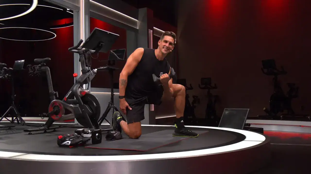 Mann mit Hantel auf einem Knie, um schnell wieder fit zu werden. Peloton-R&auml;der im Hintergrund.