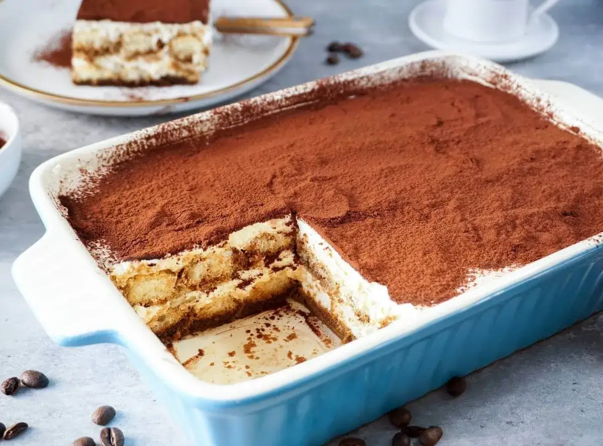 Ein cremiges Tiramisu, bestäubt mit Kakao, wartet in einer blauen Auflaufform. Ein Stück ist bereits entnommen.