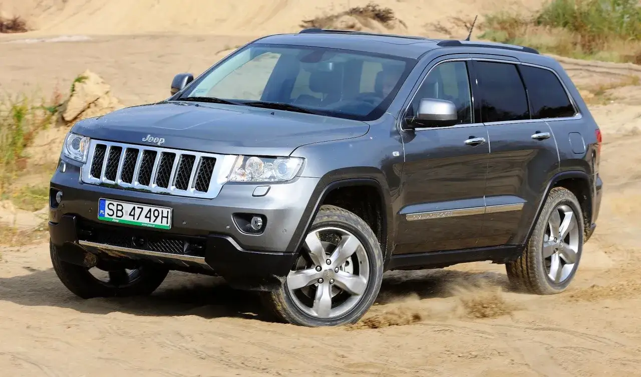 Szary Jeep Grand Cherokee WK2 pokonuje piaszczysty teren, wzbijając tumany kurzu.