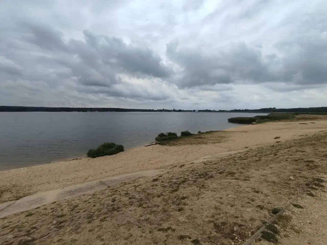Plaża nad jeziorem w Poraju, z piaszczystym brzegiem i ciemnymi chmurami na niebie.