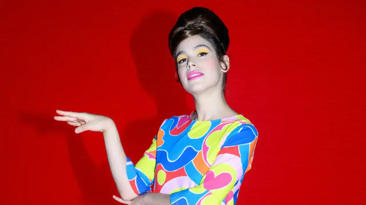 Cosima Ruiz de la Prada, con un peinado voluminoso y maquillaje vibrante, posa con un vestido de estampado psicodélico sobre fondo rojo.