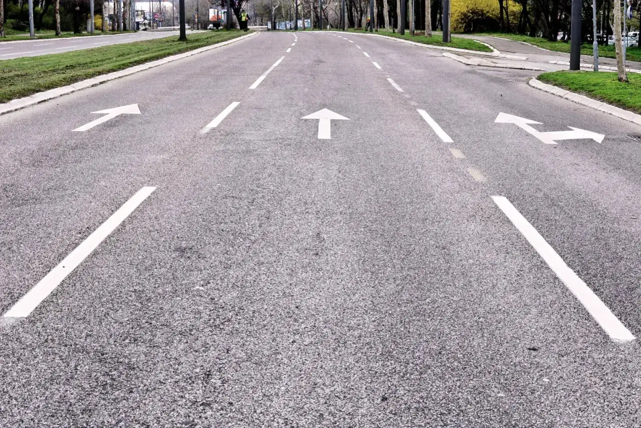 Strada con frecce bianche che indicano direzioni diverse. Una corsia di preselezione si apre per chi deve svoltare a sinistra o andare dritto.