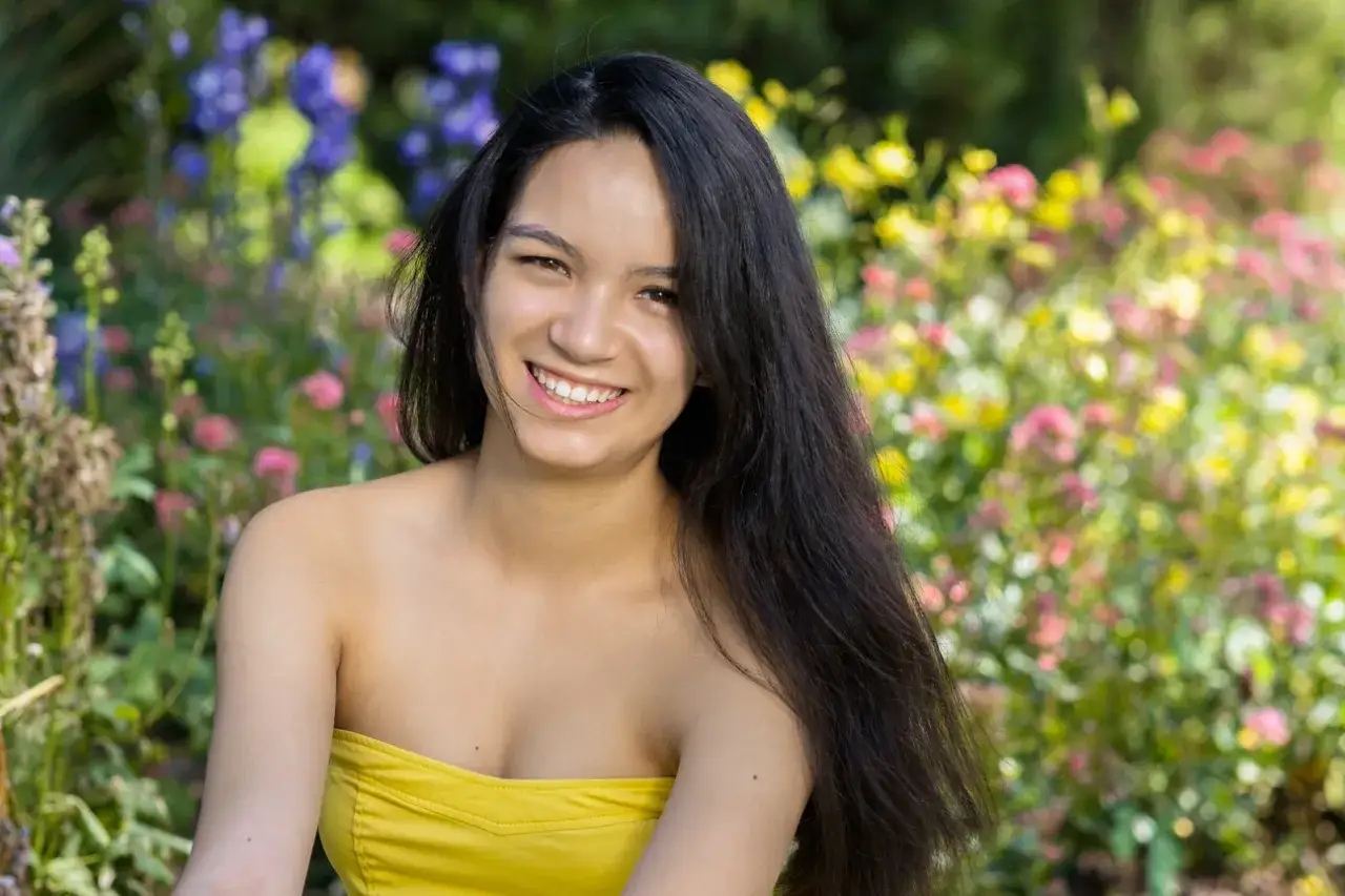 A young woman with long, dark hair smiles brightly in a garden, her yellow top contrasting with the colorful flowers. Shot with a 50mm lens at f/1.8, the background is beautifully blurred.