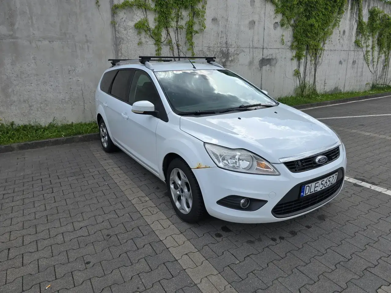 Biały Ford Focus 2 kombi z bagażnikiem dachowym stoi na parkingu wyłożonym kostką.