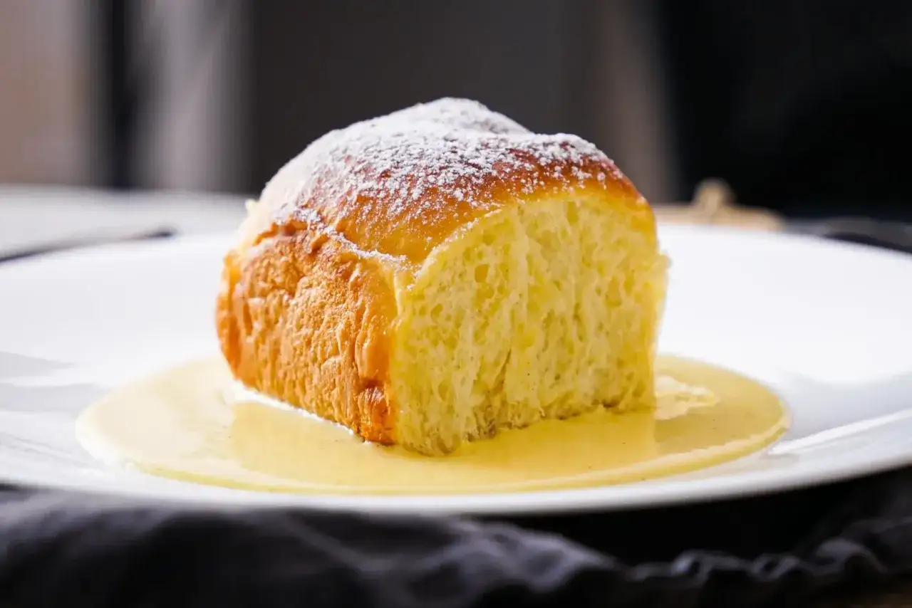Ein fluffiges Buchtel-Rezept, bestäubt mit Puderzucker, serviert in warmer Vanillesauce auf einem weißen Teller.