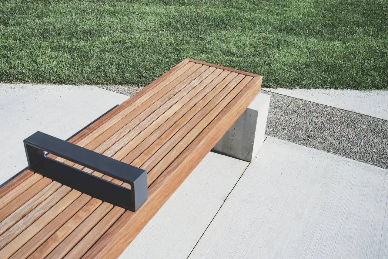 Banco moderno con asiento de madera resistente al agua, apoyabrazos met&aacute;lico y base de hormig&oacute;n, junto a c&eacute;sped y grava.