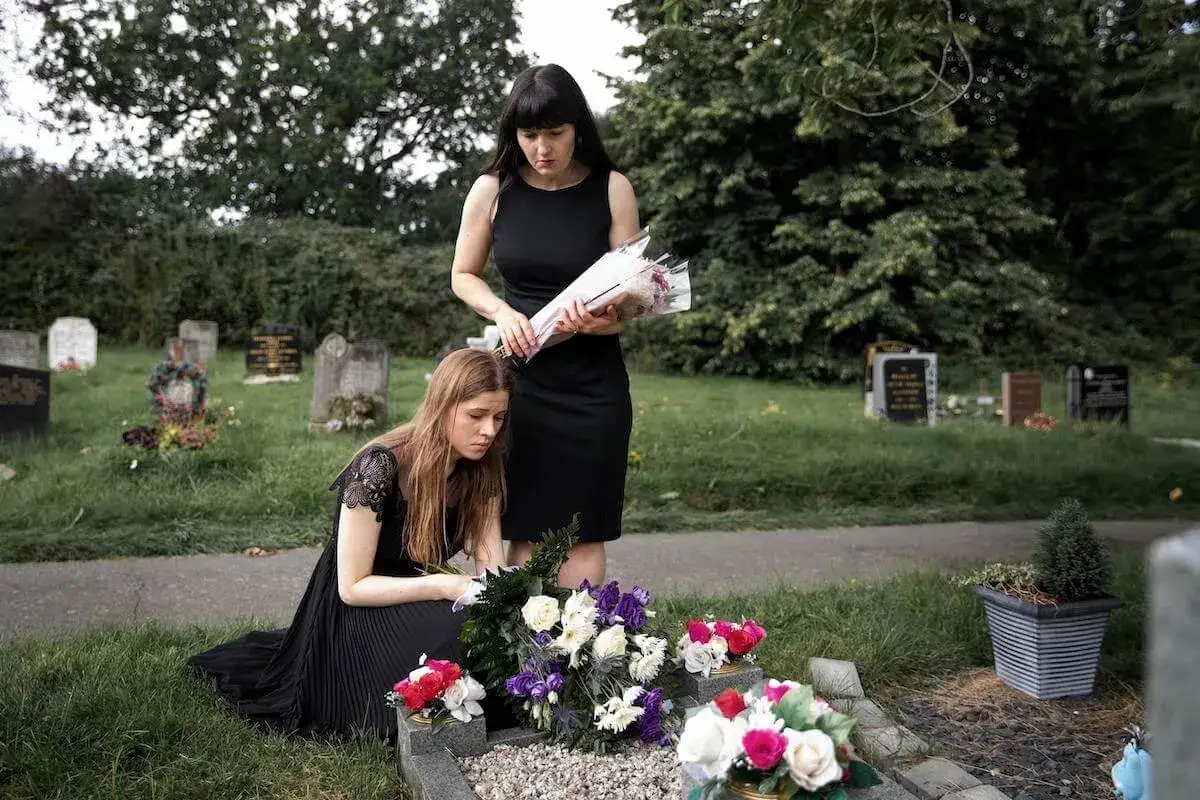 Dwie kobiety w czerni składają kwiaty na grobie. Jedna klęczy przy pomniku, druga stoi z bukietem.