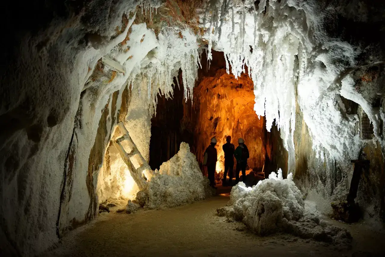 Exploradores admiran las formaciones de sal en una mina de sal, con estalactitas blancas y una luz c&aacute;lida al fondo.
